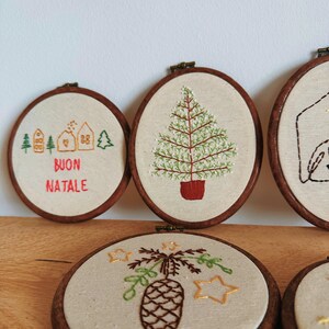 May include: Embroidered Christmas ornaments in wooden hoops. Designs include a Christmas tree, pine cone, houses, and the text "BUON NATALE". The ornaments are on a light-colored fabric background.