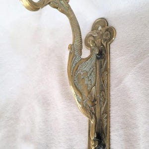 May include: A brass wall hook featuring an elongated, sculpted bird figure with detailed wing textures. The hook extends from the bird's head, and the vertical mounting plate has a decorative, scalloped edge with two circular screw holes.