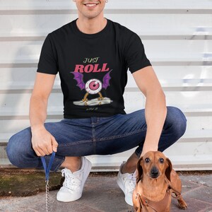 May include: Black t-shirt with the text "JUST ROLL" above a cartoon eyeball with bat wings. The person is wearing blue jeans and white sneakers. A brown dachshund dog is wearing a brown harness and is on a leash.