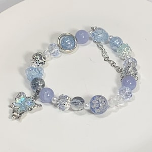 以下が含まれることがあります： 銀の蝶のチャーム、青と白のビーズ、銀のチェーンが付いた繊細なブレスレットです。