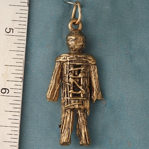 May include: Gold-toned pendant charm depicting a human figure wrapped in a woven material. The figure is standing with arms at its sides and has a detailed, textured surface.