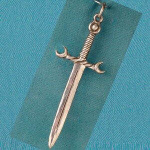 May include: A silver sword pendant with a twisted handle and crescent-shaped guards. The pendant is hanging against a blue background.