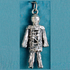 May include: A silver pendant in the shape of a stylized human figure. The figure has a textured surface and is detailed with lines and cross-hatching, giving it a unique, handcrafted appearance. The pendant hangs from a small loop.