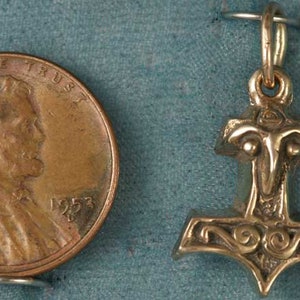May include: A gold-colored pendant in the shape of Thor's hammer, next to a U.S. penny. The pendant has intricate details and a loop for a chain. The penny shows a profile of Abraham Lincoln and the year 1953.
