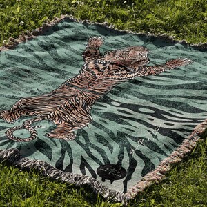 May include: A green and black woven throw blanket with a tiger design. The tiger is orange and black and is lying on its back with its paws in the air. The blanket has a fringe border.
