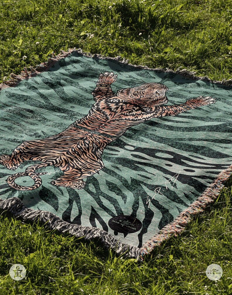 May include: A green and black woven throw blanket with a tiger design. The tiger is orange and black and is lying on its back with its paws in the air. The blanket has a fringe border.