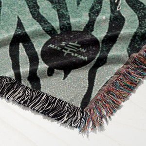 May include: A green and black woven throw blanket with a fringe border. The blanket features a repeating pattern of black stripes and a circular design with the text "Sam" in the center.