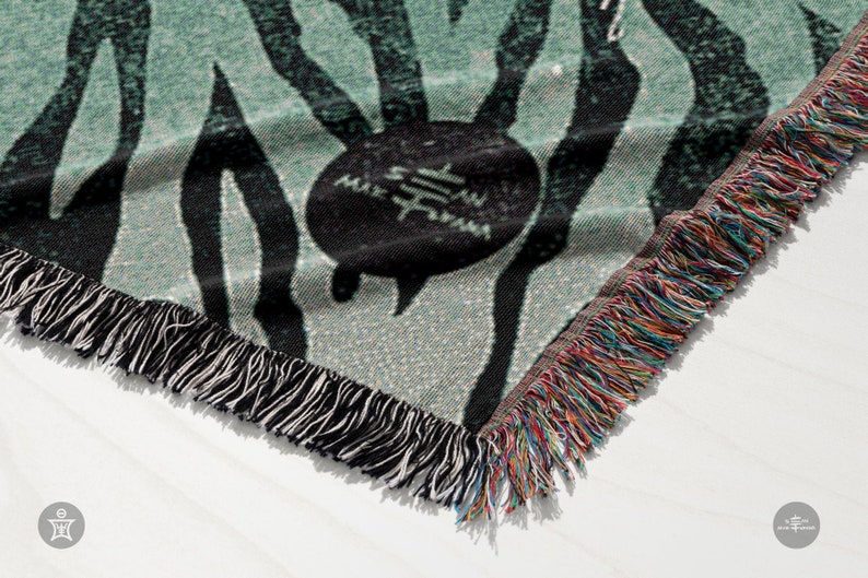 May include: A green and black woven throw blanket with a fringe border. The blanket features a repeating pattern of black stripes and a circular design with the text "Sam" in the center.