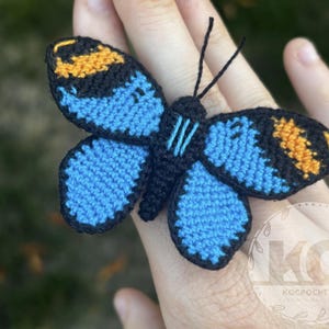 May include: A handmade crochet butterfly with blue wings, black edging, and orange and yellow accents. The butterfly has black antennae and is likely a decorative item or accessory.