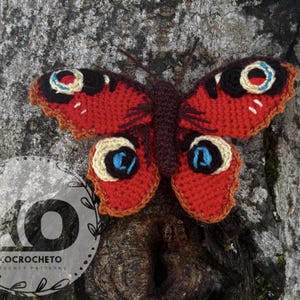 May include: A handmade crochet butterfly with red, black, and cream-colored wings, and a brown body. The butterfly is attached to a tree trunk. The wings have a detailed pattern. The logo "KOCROCHETO CROCHET PATTERNS" is visible.