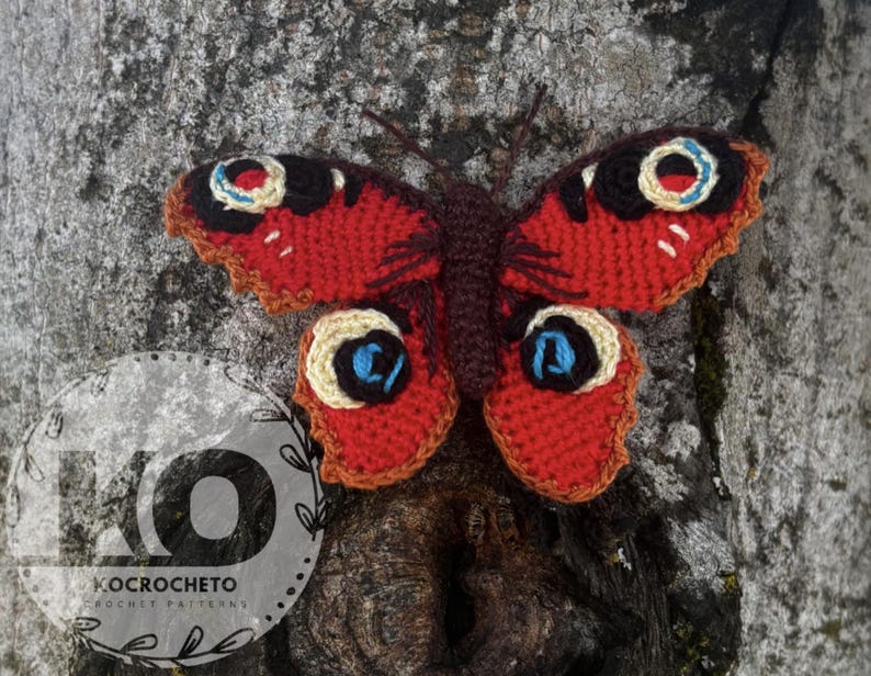 May include: A handmade crochet butterfly with red, black, and cream-colored wings, and a brown body. The butterfly is attached to a tree trunk. The wings have a detailed pattern. The logo "KOCROCHETO CROCHET PATTERNS" is visible.
