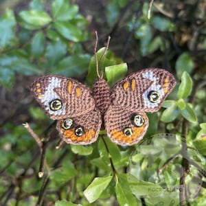 Puede incluir: Una mariposa de ganchillo hecha a mano con detalles en marrón, naranja y blanco. La mariposa está posada en una rama entre hojas verdes. Las alas tienen patrones intrincados y el cuerpo es de color marrón sólido. Un artículo decorativo.