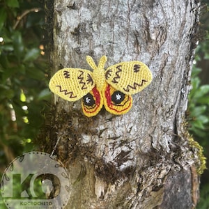Puede incluir: Una polilla de ganchillo amarilla hecha a mano con detalles marrones y rojos, adherida a un tronco de árbol. La polilla presenta intrincados diseños de alas y un diseño delicado. La imagen resalta la textura del ganchillo y el entorno natural.