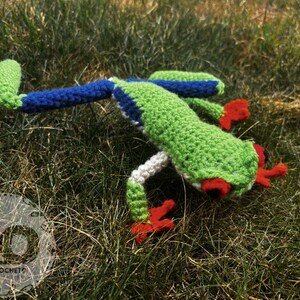 May include: A handmade crochet frog in a grassy setting. The frog features a green body with blue and white accents, red feet, and red eyes. The frog is lying on the grass.