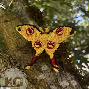 May include: A handmade, yellow crochet moth with red and black accents. The moth features detailed wing patterns and is displayed against a natural, tree bark background. The wings have red and black details. The moth is a decorative item.