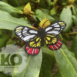 Op de afbeelding: Een handgemaakte gehaakte vlinder met zwarte, witte, gele en rode details, rustend op een groen blad. De vleugels van de vlinder hebben ingewikkelde patronen. De tekst "KOCROCHETO CROCHET PATTERN" is inbegrepen.