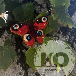 May include: A handmade crochet butterfly with red and brown wings, detailed with blue and cream eye-like markings. The butterfly is resting on green leaves and a tree trunk. The image includes the text "KOCROCHETC CROCHET PATTERNS".