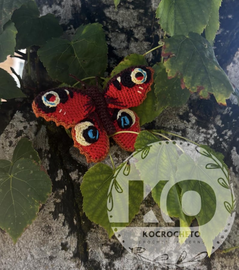 May include: A handmade crochet butterfly with red and brown wings, detailed with blue and cream eye-like markings. The butterfly is resting on green leaves and a tree trunk. The image includes the text "KOCROCHETC CROCHET PATTERNS".