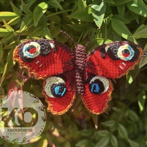 May include: A handmade crochet butterfly with vibrant red wings, detailed with black, white, and blue eye-like markings. The butterfly is set against a backdrop of green foliage, showcasing the intricate craftsmanship of the textile art.
