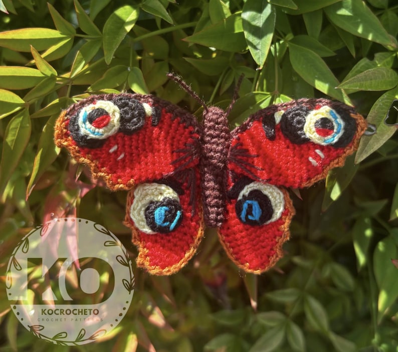 May include: A handmade crochet butterfly with vibrant red wings, detailed with black, white, and blue eye-like markings. The butterfly is set against a backdrop of green foliage, showcasing the intricate craftsmanship of the textile art.