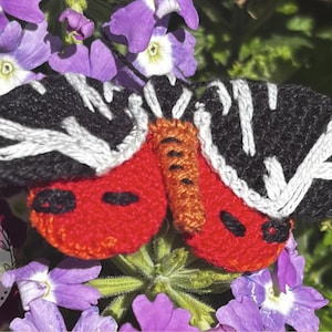 May include: A handmade butterfly brooch with black and white embroidered wings and red body. The brooch is set against a backdrop of purple flowers and green foliage, creating a vibrant and natural setting. This unique accessory adds a touch of nature-inspired charm.