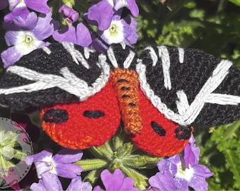 Puede incluir: Broche de mariposa hecho a mano con alas bordadas en negro y blanco y cuerpo rojo. El broche se encuentra sobre un fondo de flores moradas y follaje verde, creando un entorno vibrante y natural. Este accesorio único añade un toque de encanto inspirado en la naturaleza.