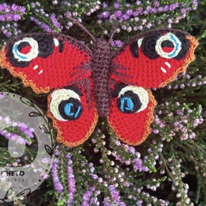 May include: A handmade crochet butterfly with red wings, detailed with black, white, and blue eye-like markings. The butterfly is set against a backdrop of purple flowers and green foliage, creating a vibrant contrast.
