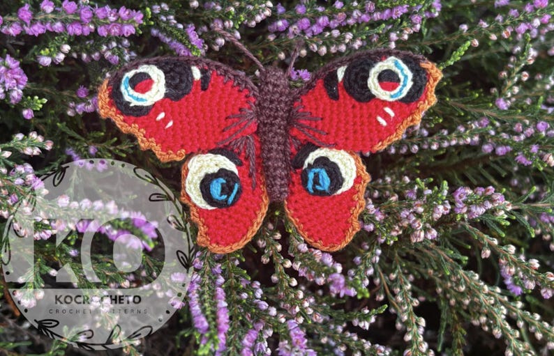 May include: A handmade crochet butterfly with red wings, detailed with black, white, and blue eye-like markings. The butterfly is set against a backdrop of purple flowers and green foliage, creating a vibrant contrast.