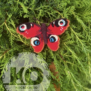 May include: A handmade crochet butterfly with red wings, detailed with white, blue, and black eye-like markings. The butterfly is set against a backdrop of green foliage, showcasing its vibrant colors and intricate design.