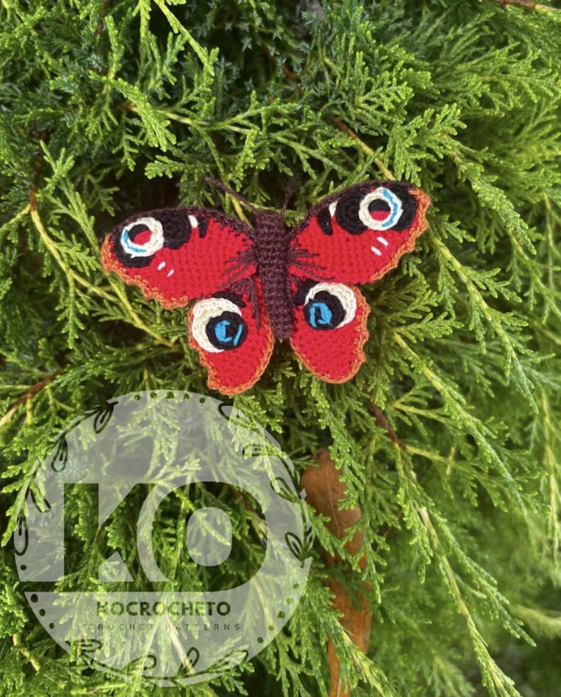 May include: A handmade crochet butterfly with red wings, detailed with white, blue, and black eye-like markings. The butterfly is set against a backdrop of green foliage, showcasing its vibrant colors and intricate design.