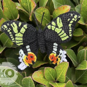 Op de afbeelding: Een handgemaakte gehaakte vlinder met zwarte vleugels met limoengroene en witte accenten. De onderste vleugels van de vlinder hebben oranje en witte details. De vlinder staat tegen een achtergrond van groene bladeren.