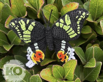 May include: A handmade crochet butterfly with black wings featuring lime green and white accents. The butterfly's lower wings have orange and white details. The butterfly is set against a backdrop of green leaves.