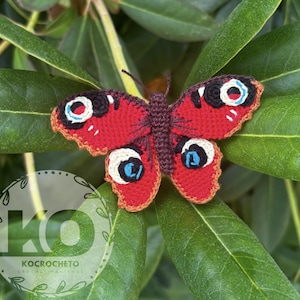 May include: A handmade crochet butterfly with red wings, featuring black, white, and blue eye-like markings. The butterfly is set against a backdrop of green leaves. The wings have a scalloped edge. The butterfly is a craft item.