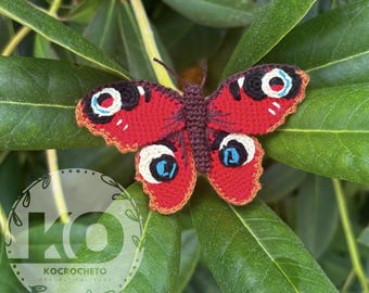 May include: A handmade crochet butterfly with red wings, featuring black, white, and blue eye-like markings. The butterfly is set against a backdrop of green leaves. The wings have a scalloped edge. The butterfly is a craft item.