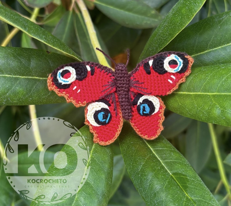 May include: A handmade crochet butterfly with red wings, featuring black, white, and blue eye-like markings. The butterfly is set against a backdrop of green leaves. The wings have a scalloped edge. The butterfly is a craft item.