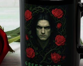 May include: A black ceramic mug featuring a graphic of a man with dark hair surrounded by red roses and green vines, with the text "GREEN MAN" printed in green at the bottom. A single red rose with a green stem is positioned next to the mug.