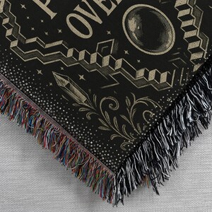 May include: Black throw blanket with a detailed design featuring geometric shapes, a crystal, and the word "OVER". The blanket has a fringe border with multicolored threads on one side and white threads on the other.