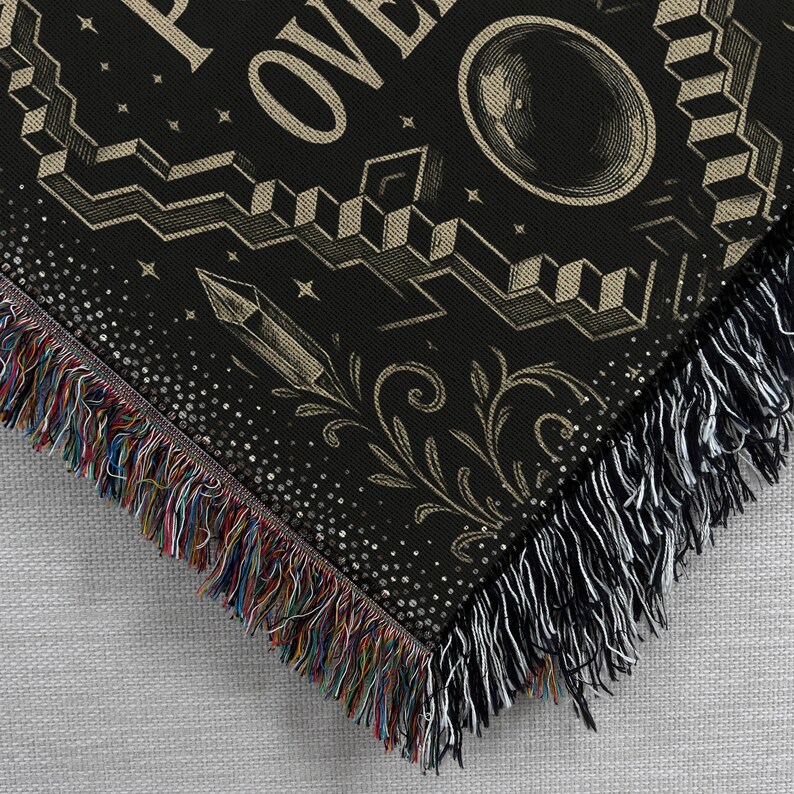 May include: Black throw blanket with a detailed design featuring geometric shapes, a crystal, and the word "OVER". The blanket has a fringe border with multicolored threads on one side and white threads on the other.