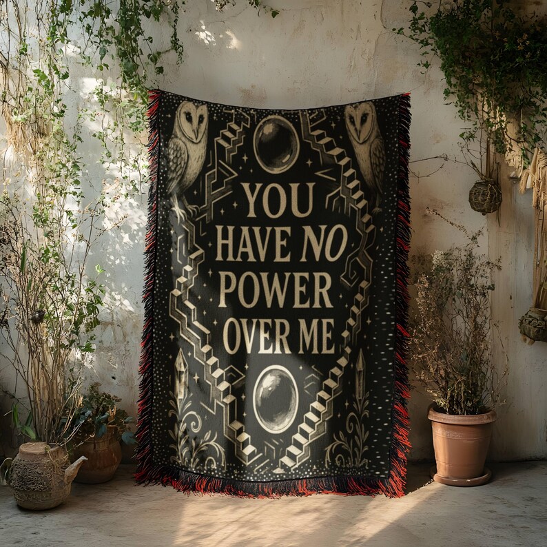 May include: Black throw blanket with the text "YOU HAVE NO POWER OVER ME" in white. The blanket features owl illustrations and geometric designs. It has red fringe along the edges, adding a decorative touch. The blanket is displayed against a textured wall.