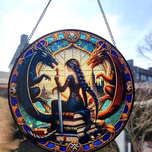 May include: Round stained glass window art featuring a woman with a sword, seated between a black and orange dragon. The woman has long dark hair and is surrounded by books. The window has a blue and gold border and is suspended by a chain.