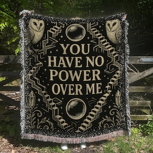 May include: Black woven throw blanket with the text "YOU HAVE NO POWER OVER ME". The design includes owls, staircases, crystals, and orbs in beige and white. The blanket has a fringed edge.