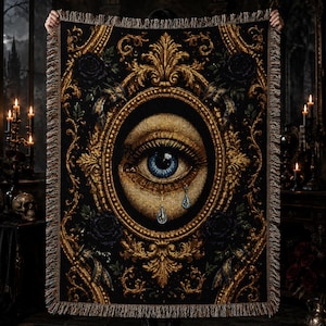 Könnte beinhalten: Eine rechteckige Webdecke mit einem zentralen blauen Auge und zwei Tränen, eingefasst in einen verzierten ovalen Goldrahmen. Der Hintergrund ist schwarz mit komplexen goldenen Ornamenten, Schnörkeln und dunklen Blumenmotiven, mit Fransen.