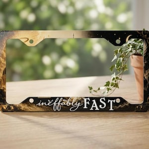 May include: A rectangular license plate frame featuring a dark celestial design with gold clouds, stars, and a vintage car silhouette. The bottom section displays the white text "ineffably FAST" in a combination of script and bold serif fonts.