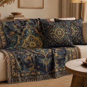 Op de afbeelding: Een geweven plaid en twee bijpassende vierkante kussens met een complex bloemrijk damastpatroon in donkerblauw, groenblauw en goud. De plaid heeft een franjerand aan de onderkant en de kussens hebben een centraal medaillonontwerp.
