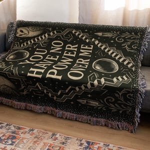 May include: Black woven throw blanket with the text "YOU HAVE NO POWER OVER ME" in white. The blanket features owl illustrations, geometric shapes, and fringe trim. The blanket is draped over a gray sofa.