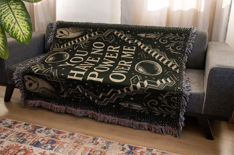 May include: Black woven throw blanket with the text "YOU HAVE NO POWER OVER ME" in white. The blanket features owl illustrations, geometric shapes, and fringe trim. The blanket is draped over a gray sofa.