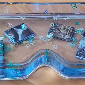 May include: A clear resin tray with a blue tint, containing miniature book replicas and scattered letter-shaped confetti. The tray has a wavy design in the center and is filled with small, detailed book covers.