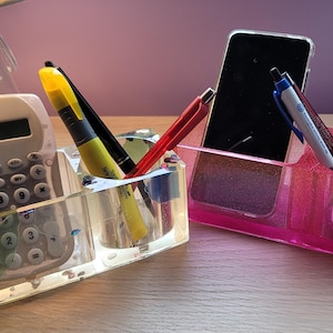 May include: Two clear plastic desk organizers with compartments for pens, pencils, and a phone. One organizer is clear and the other is pink with glitter.