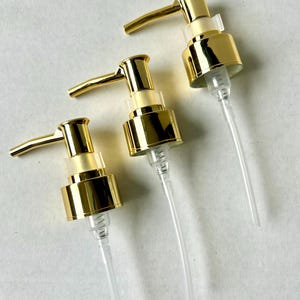 May include: Three gold-colored pump dispensers with clear plastic tubes. The pumps have a shiny, metallic finish and a lever for dispensing. The clear tubes extend downwards, suggesting they are designed for use with bottles or containers.