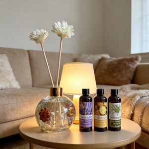 Op de afbeelding: Een heldere glazen diffuser met bloemaccenten en twee witte bloemstelen staat op een ronde tafel. Drie essentiële olieflessen met de labels "Lavendel", "Ananas" en "Citroengras" staan naast de diffuser. Een brandende lamp en een beige bank zijn op de achtergrond.